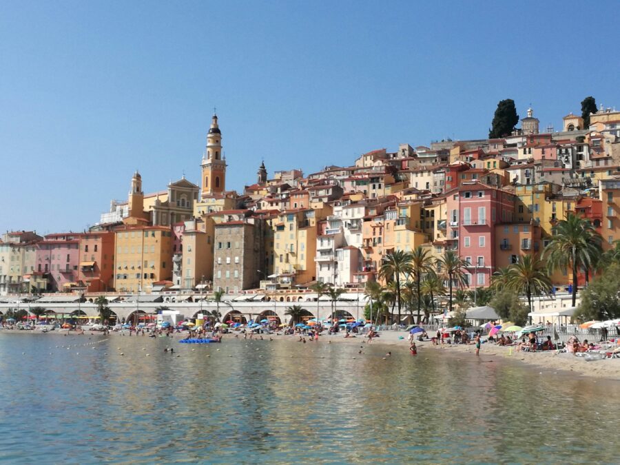 Menton, French Riviera beach with pastel hillside buildings, palm trees, and bell tower.