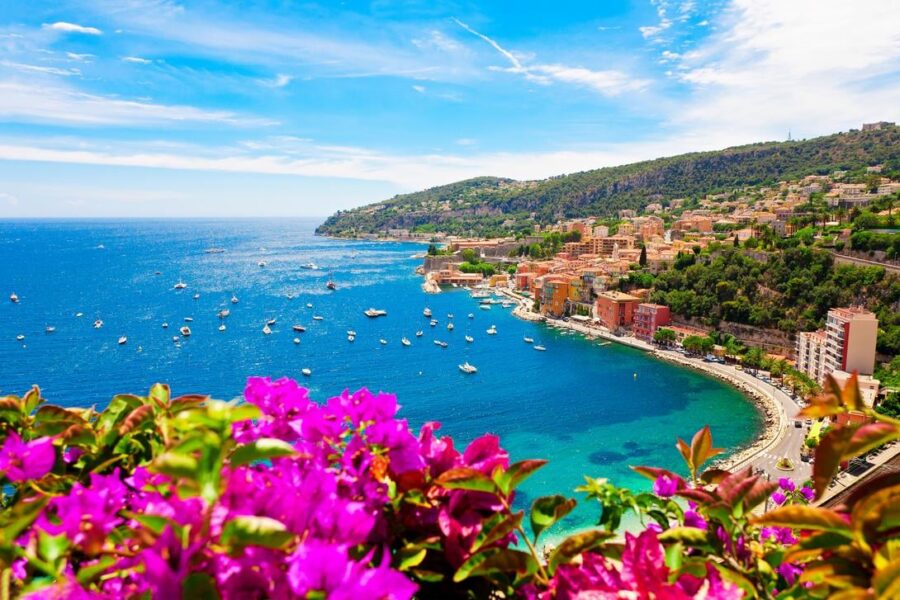 Mediterranean bay scene with vibrant flowers, boats, beaches, and hillside architecture in sunlight.