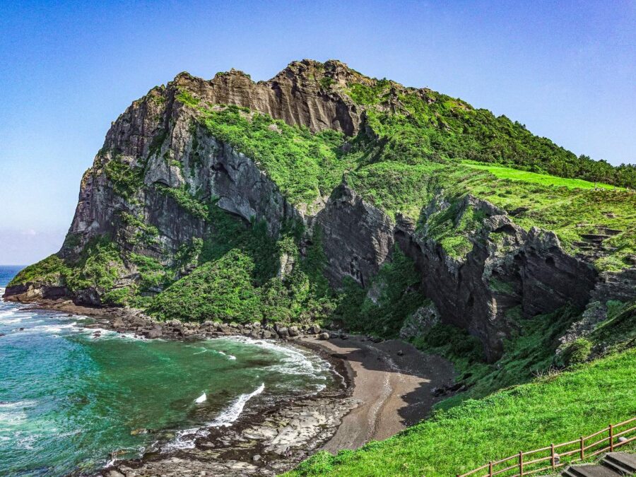 Dramatic Seongsan Ilchulbong volcanic cliff with lush green slopes above turquoise Jeju Island bay.