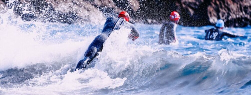Adventure Triathlons scene with wetsuit-clad individuals against crashing waves and rugged rock formations.