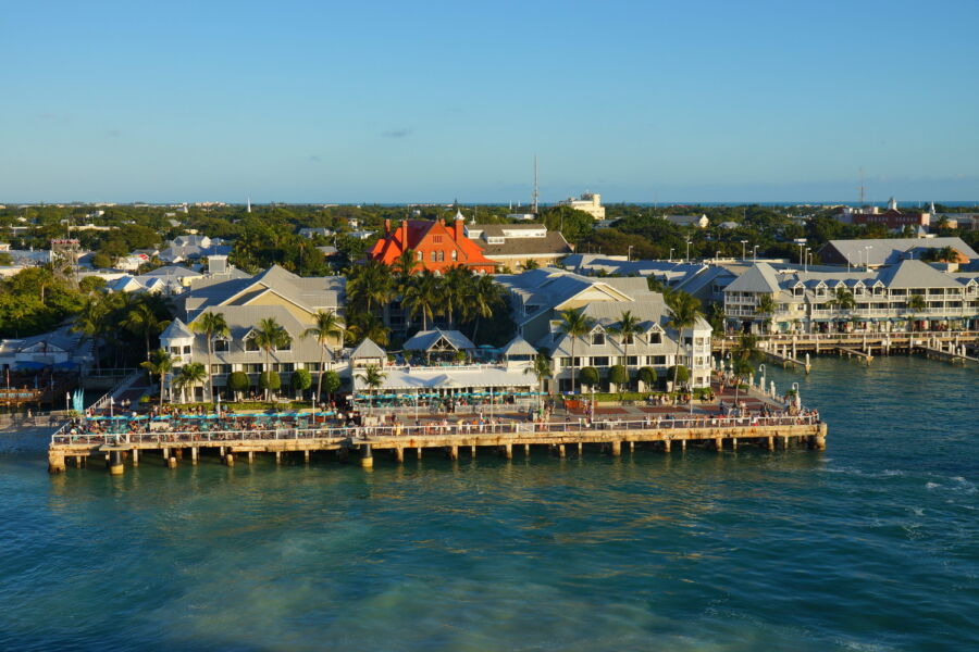 Tropical waterfront scene with pier, vibrant buildings, palm trees, and sparkling blue water.