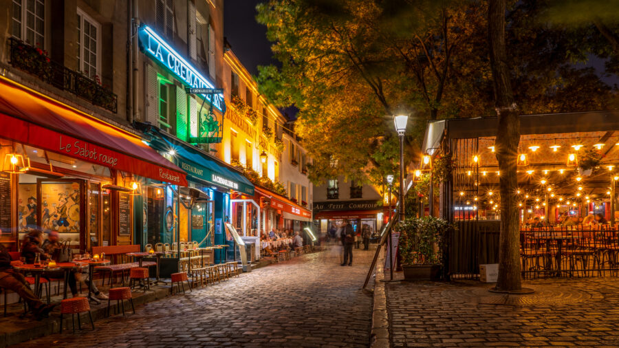Cobblestone Paris cafe street at night with outdoor dining, neon signs, and warm lights.