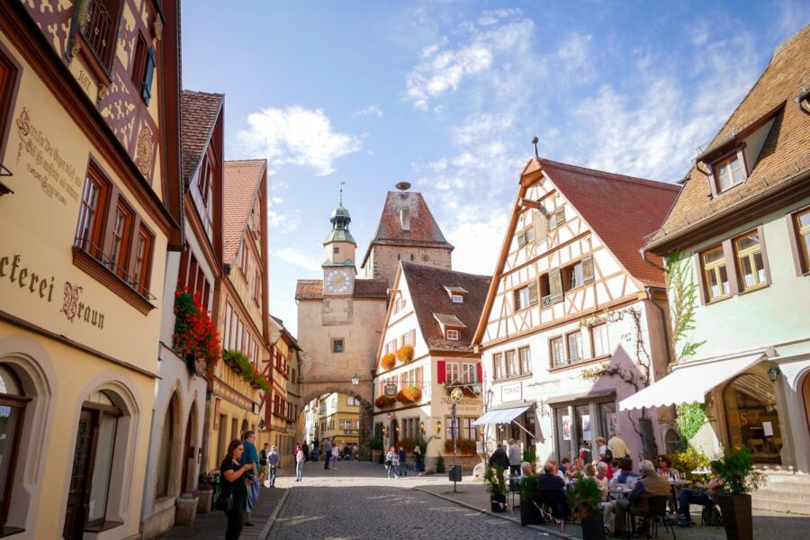 Quaint historic town scene with cobblestone streets, clock tower, half-timbered buildings, cafes, and shops.