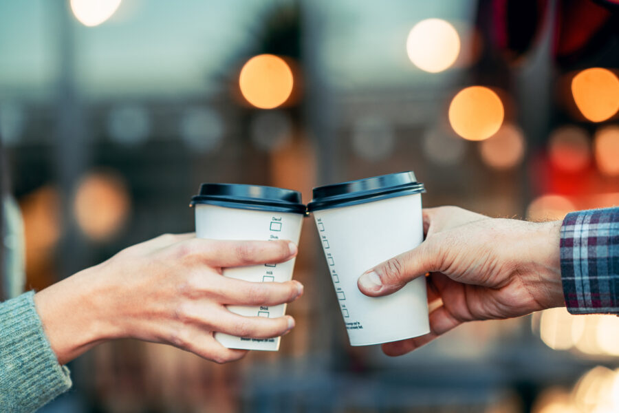 Two hands hold white coffee cups with black lids against a blurred background of warm, glowing lights, conveying a cozy and cheerful atmosphere.