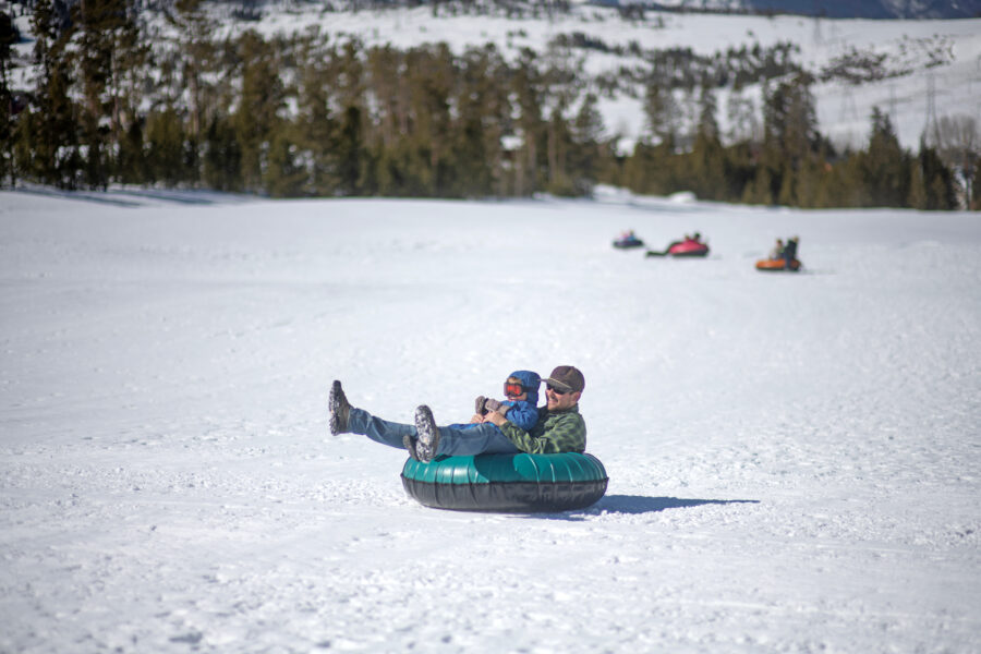 Father and son joyfully snow tube down a snowy hill in Colorado, surrounded by winter scenery