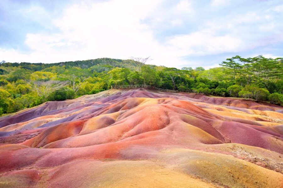 Colorful Chamarel earth dunes landscape with layered rainbow soil hills, green forest, and cloudy sky.