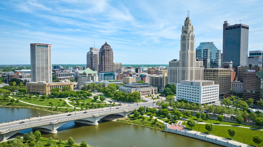 Aerial view of downtown Columbus, Ohio, showcasing skyscrapers, a green area, and a bridge with vehicles driving across 
