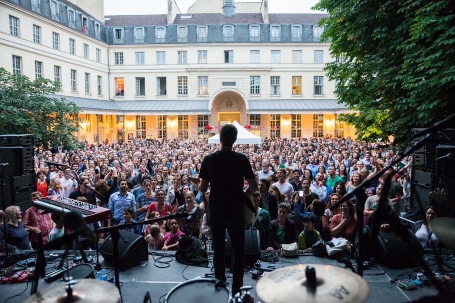 Outdoor concert in historic courtyard with musician, diverse crowd, vibrant atmosphere, and lush greenery.