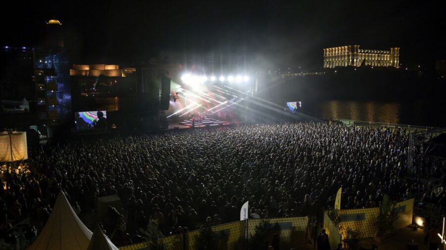 Nighttime concert scene with vibrant crowd, colorful lights, and picturesque urban backdrop.