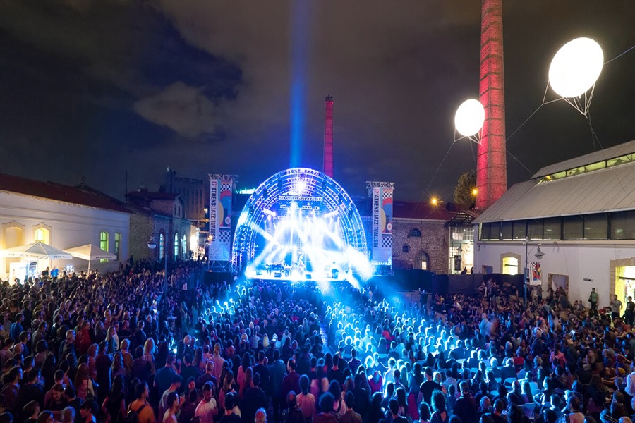 Night concert with blue-lit stage, diverse crowd, and industrial backdrop at Athens Jazz Festival.