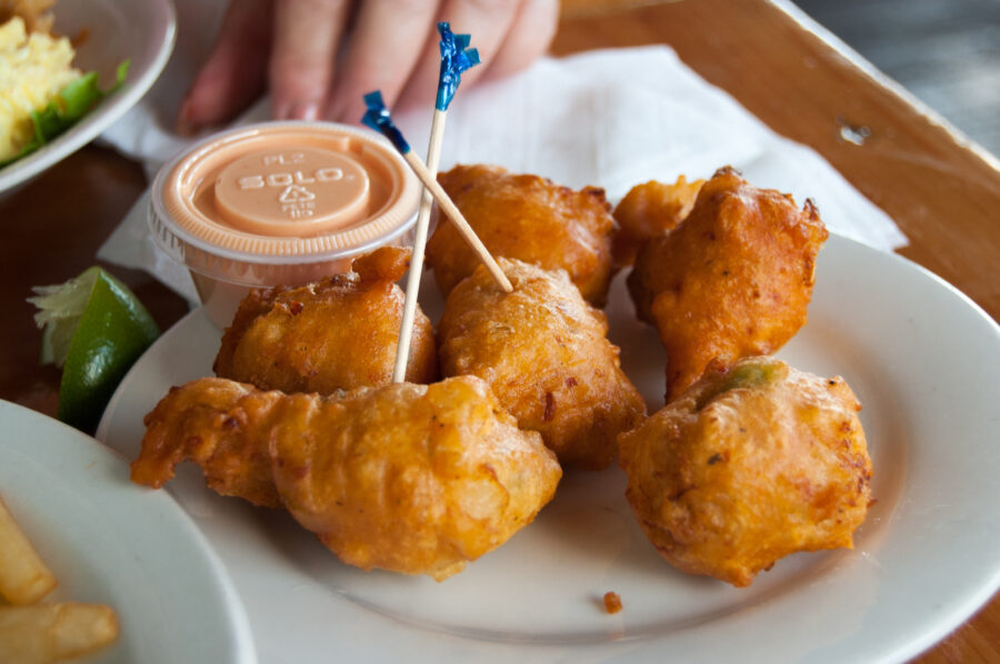 Plate of golden, crispy conch fritters with toothpicks, alongside a small container of dipping sauce and a lime wedge. Casual dining setting.