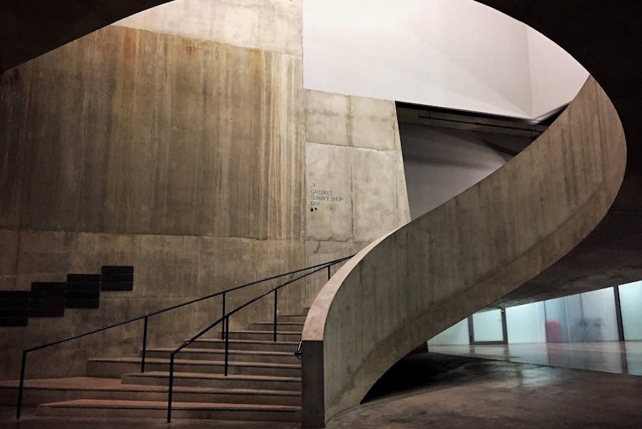Brutalist concrete museum interior with sweeping curved ramp and stepped staircase.