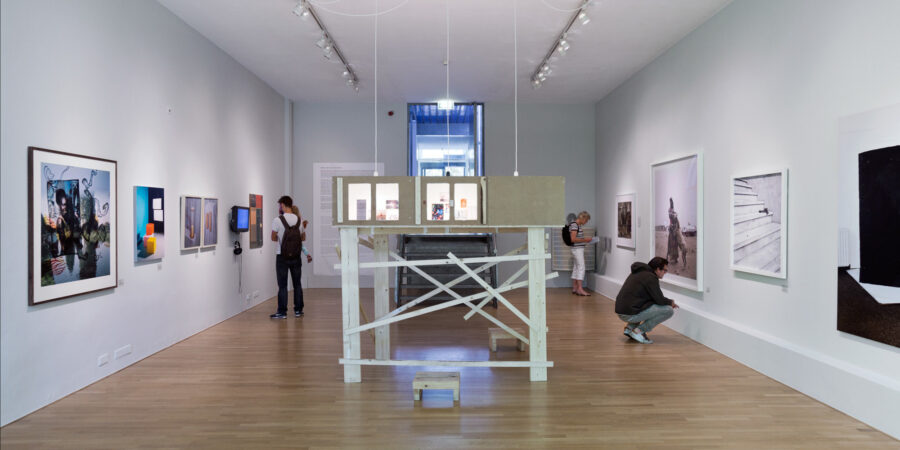 Contemporary art gallery interior with photography exhibition and central wooden installation viewed by visitors.