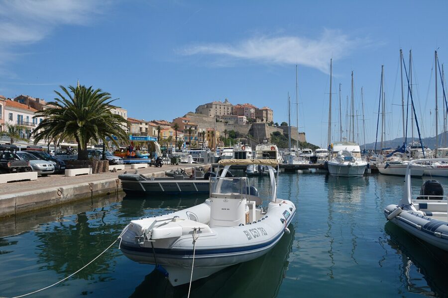Boat docked in Calvi Port, Corsica, with a historic castle rising in the background against a clear blue sky.