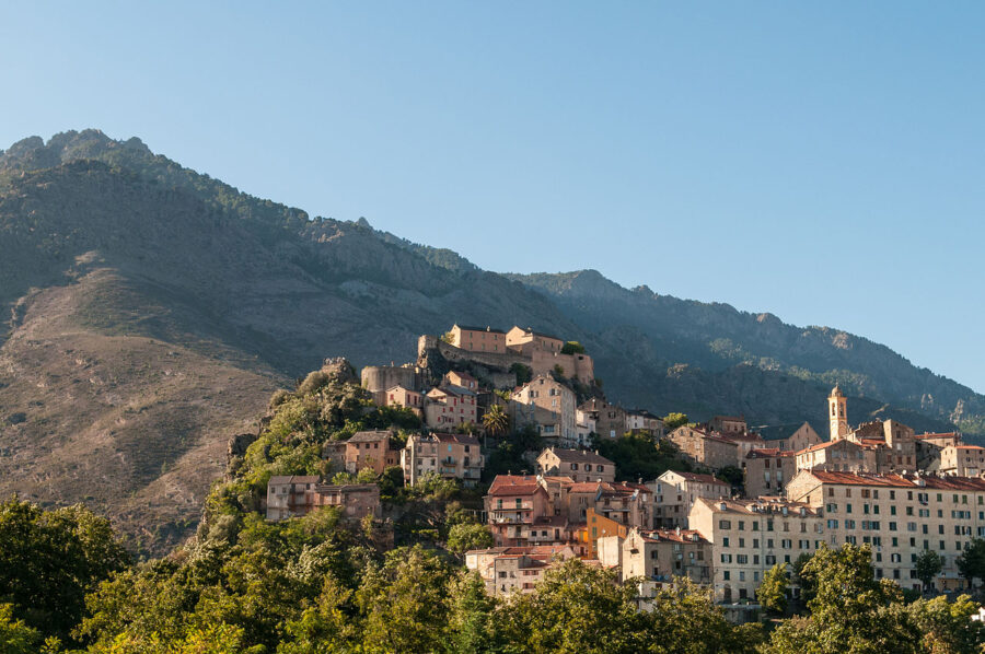 Panoramic view of Corte, Corsica, featuring a small town atop a hill with majestic mountains in the background.