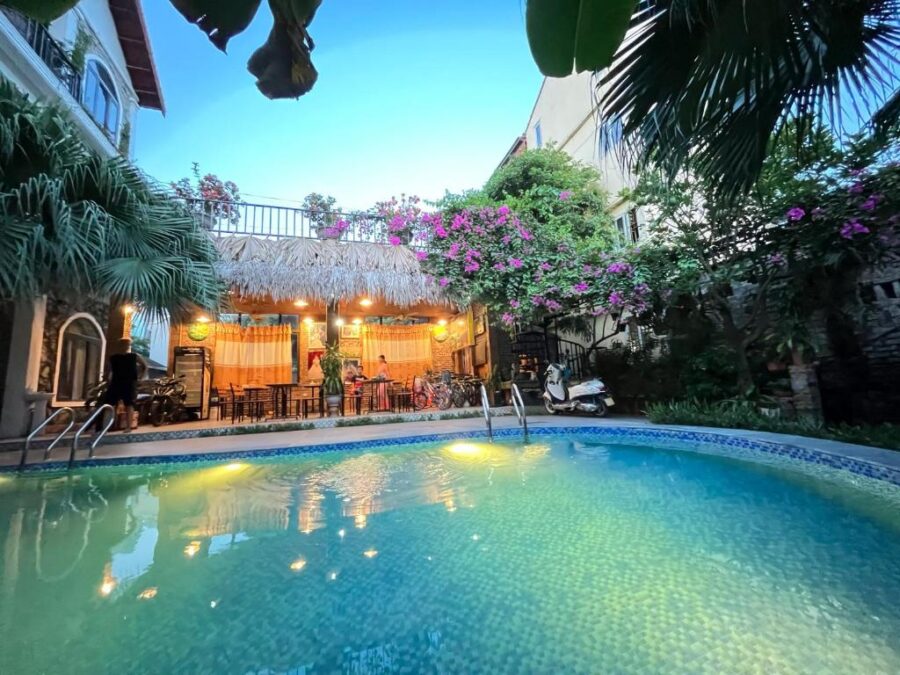Tropical Tam Coc guesthouse courtyard with illuminated pool, thatched rooftop restaurant and lush flowering garden.