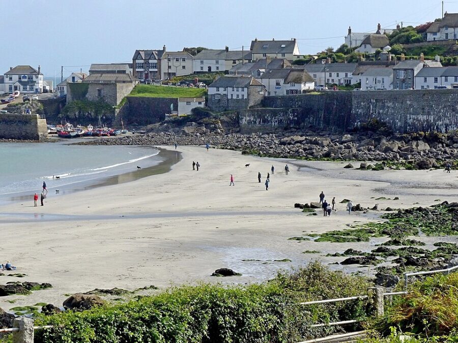 People stroll along a beach in Coverack, Lizard Peninsula, with residential houses lining the background.