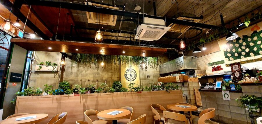 Cozy modern cafe with wooden tables, indoor plants, industrial ceiling, and order counter.