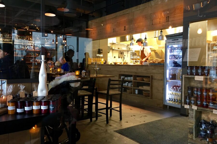 Cozy cafe with open kitchen, wood interior, display jars, bar seating, and refrigerated desserts.