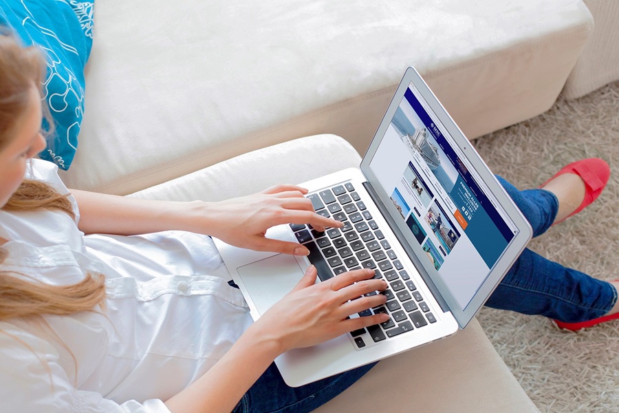 Young woman researching travel deals on laptop, sitting on a cozy couch with a blue cushion.