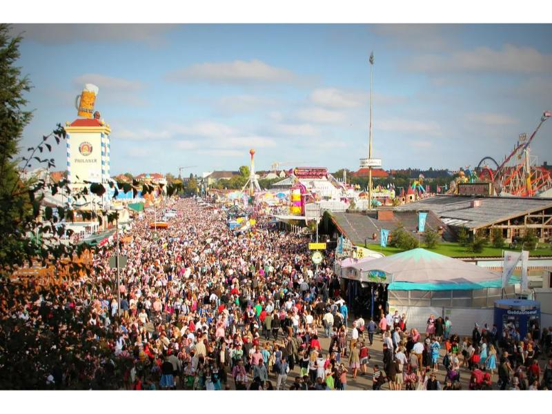 Crowded festival avenue with beer tents, amusement rides, and bustling fairground activity.