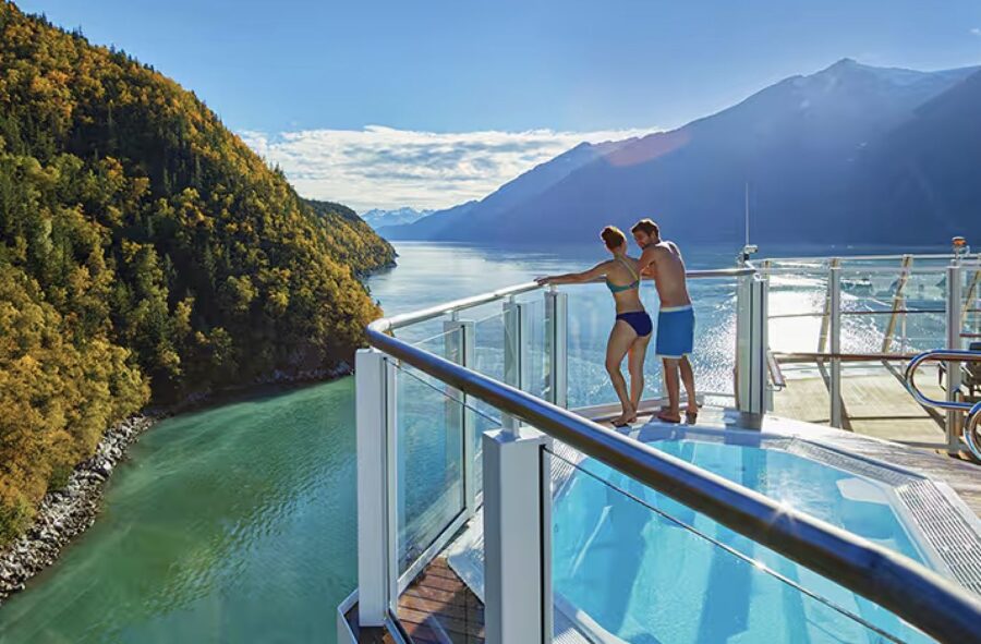 Couple enjoys romantic cruise with scenic bay, mountains, and hot tub under clear blue sky.