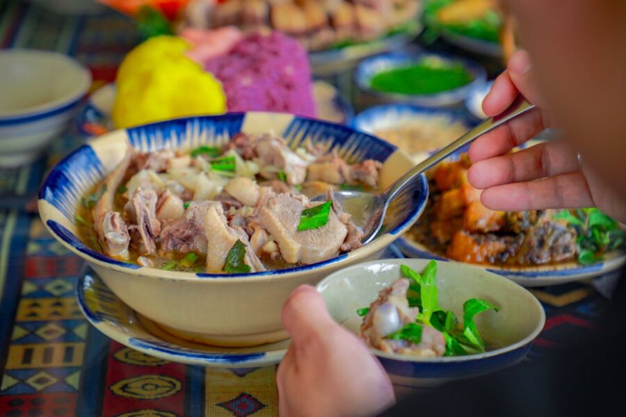 Communal dining scene with vibrant soup, meat, herbs, and patterned tablecloth.