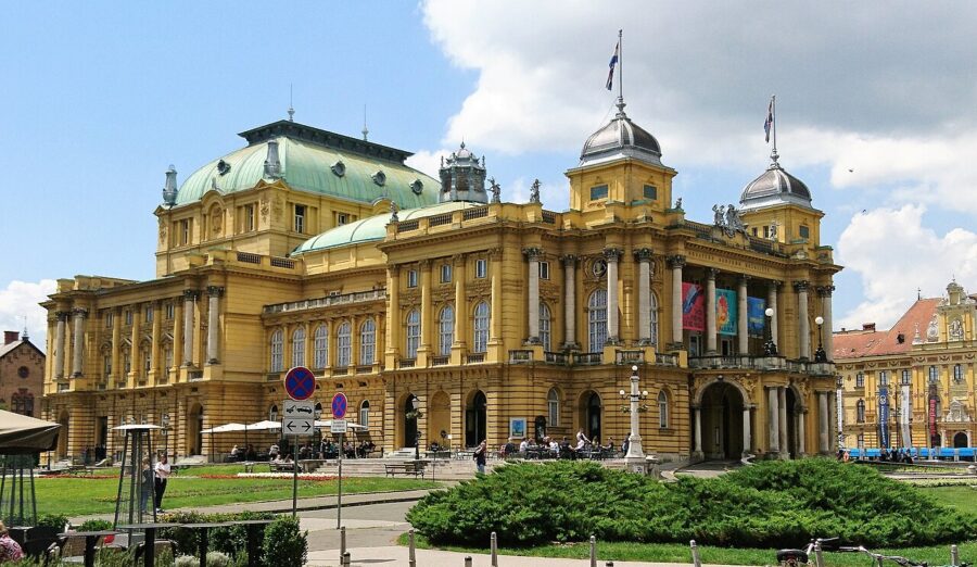 Grand Baroque-Neoclassical Croatian National Theater with vibrant yellow facade and green dome, lively surroundings.