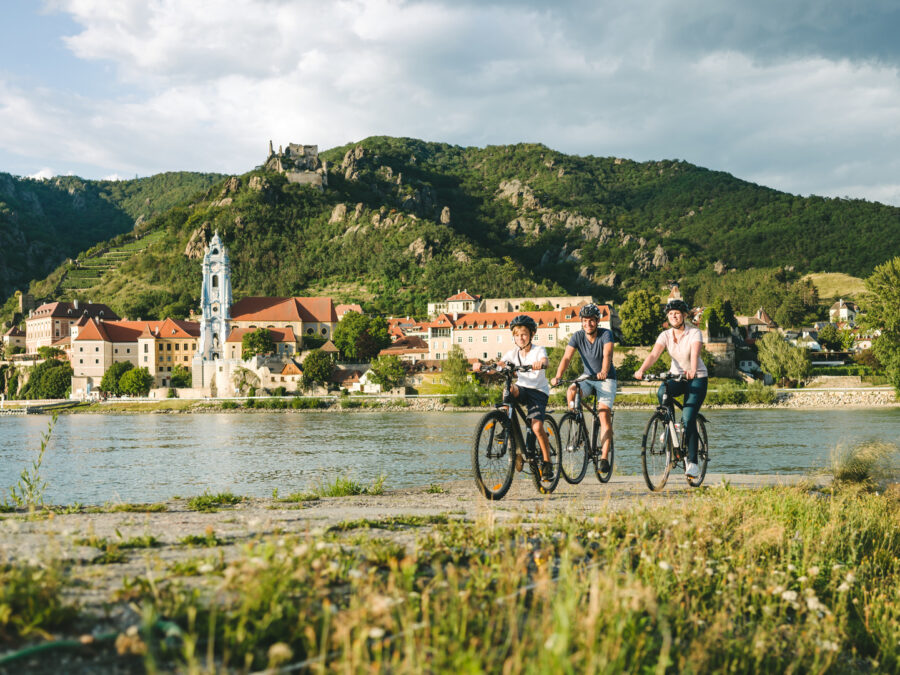 Cyclists on Danube River bike trail passing Austrian riverside village, vineyards and hilltop castle.