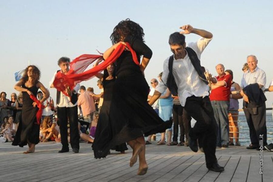 Outdoor traditional dance performance on boardwalk with audience, vibrant outfits, and scenic backdrop.