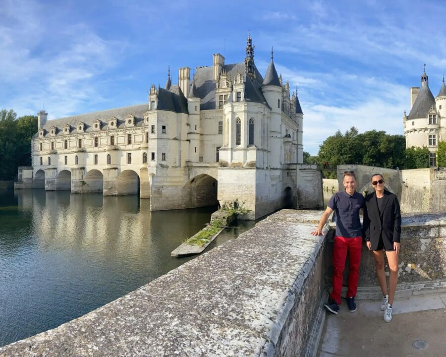 Man and woman stand on a stone bridge, smiling in front of the elegant Château de Chenonceau, which arches over a river under a clear blue sky.