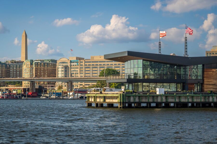 Contemporary riverside building with glass facades, urban skyline, boats, and a historic monument.