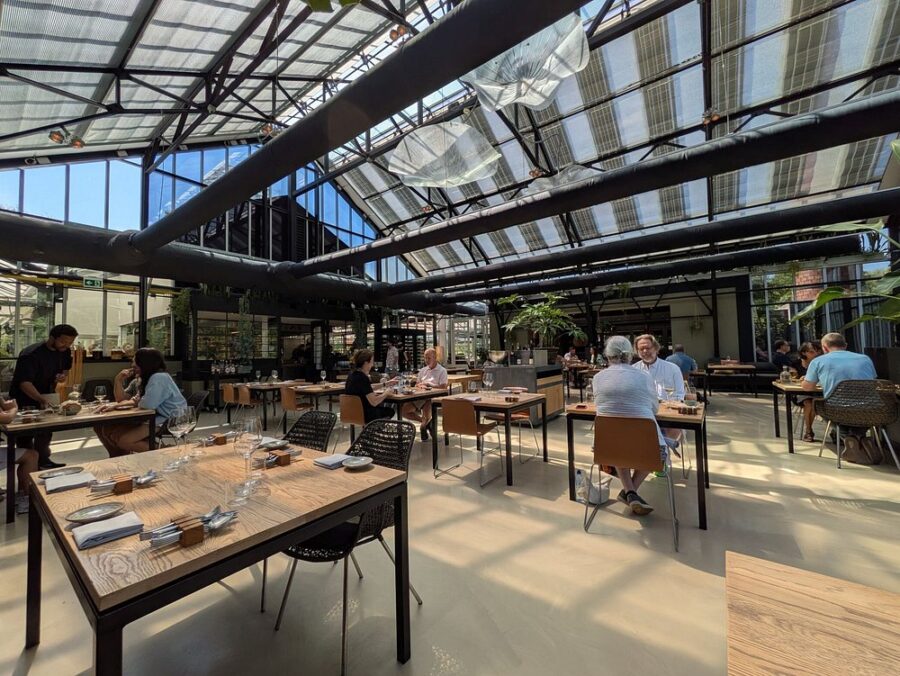 Interior of De Kas Amsterdam, featuring diners at tables beneath a spacious glass roof.