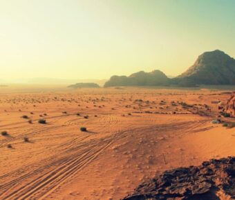 Golden desert landscape with 4x4 tire tracks, rocky cliffs, remote camp and sunset light.