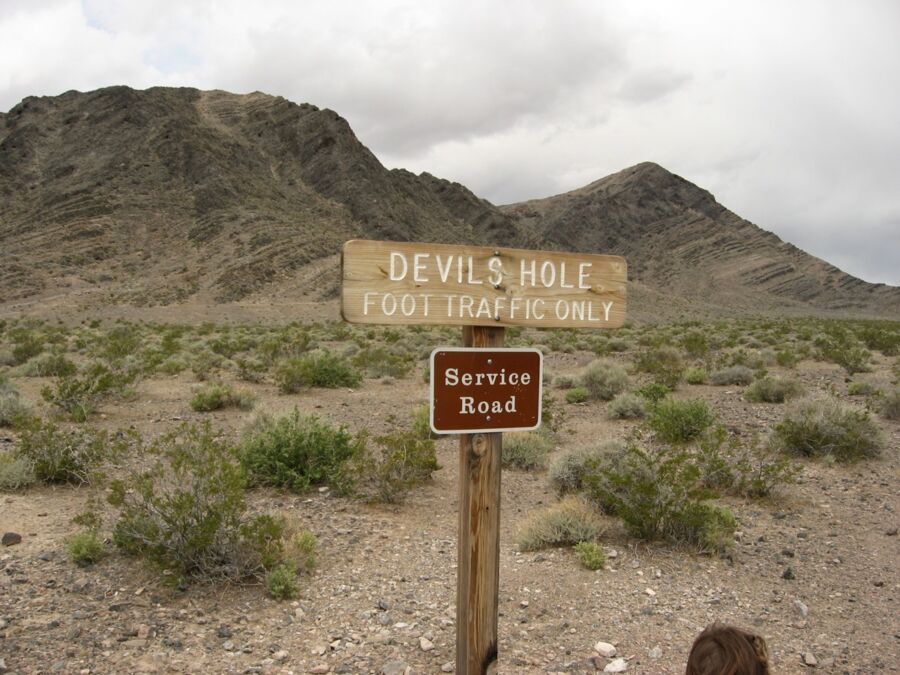 Desert landscape with Devils Hole signs, rugged terrain, mountains, and cloudy sky.