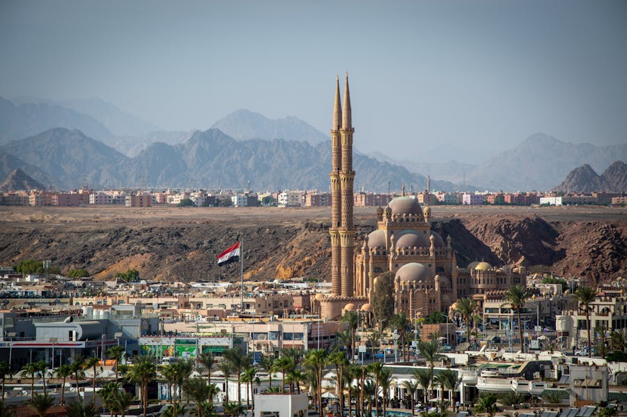 Grand desert mosque with twin minarets, city buildings, mountains, and national flag in Egypt.