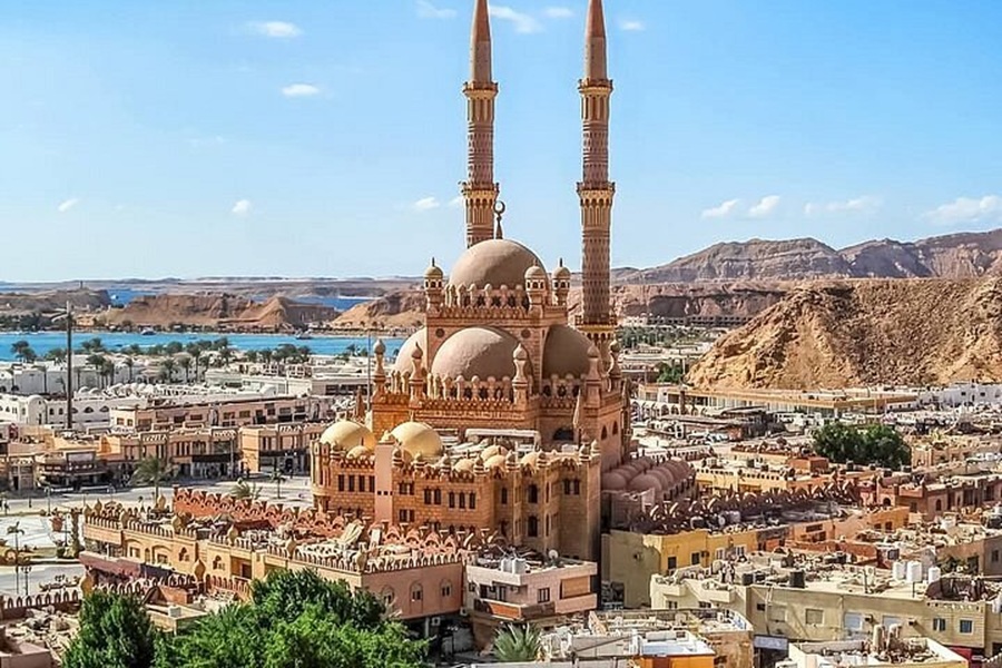 Desert cityscape with grand Islamic mosque, tall minarets, and rugged mountains near blue sea.
