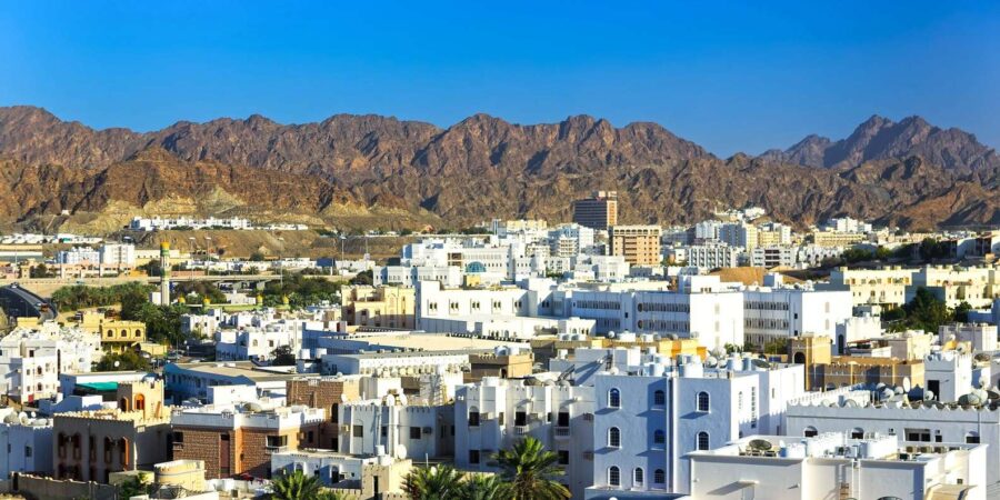 Old Muscat cityscape with white buildings, mosque minaret and rugged desert mountains under blue sky.