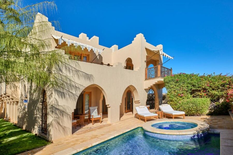 Luxury desert villa with pool, Middle Eastern architecture, sun loungers, lush garden, and clear blue sky.