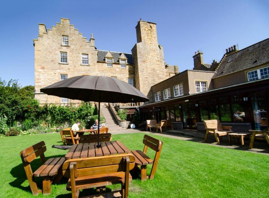 Dornoch Castle Hotel, a large stone structure, showcases outdoor wooden tables and a large umbrella for shade.