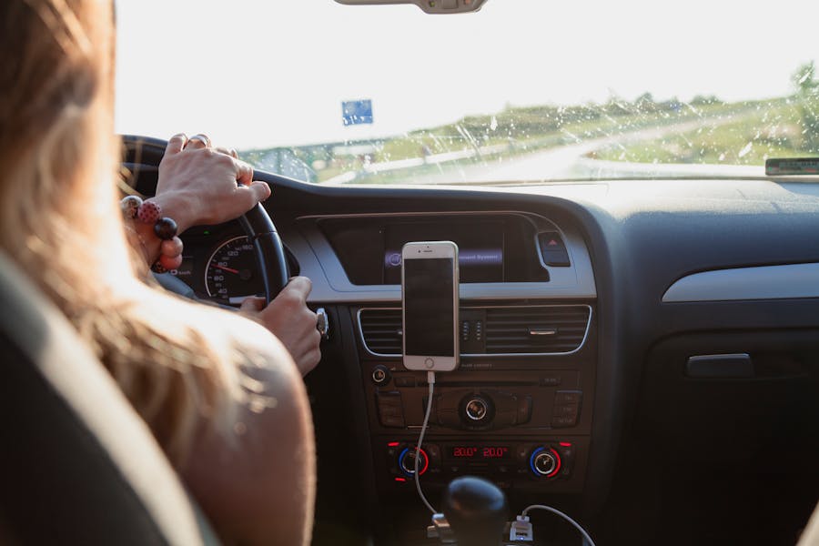 Driver hands on steering wheel, dashboard with phone mount and climate controls, road at sunset.