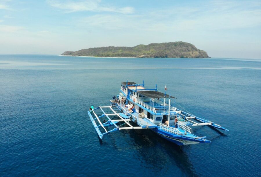 Aerial drone photo of a blue outrigger passenger boat near a forested island.