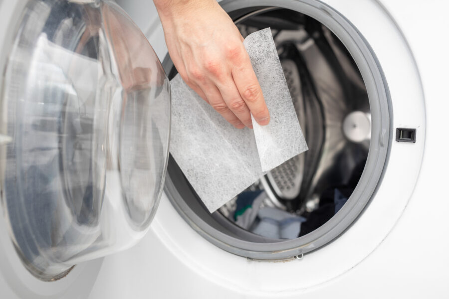 Hand places a dryer sheet into an open front-loading washing machine filled with clothes. The scene is bright and suggests a household chore.