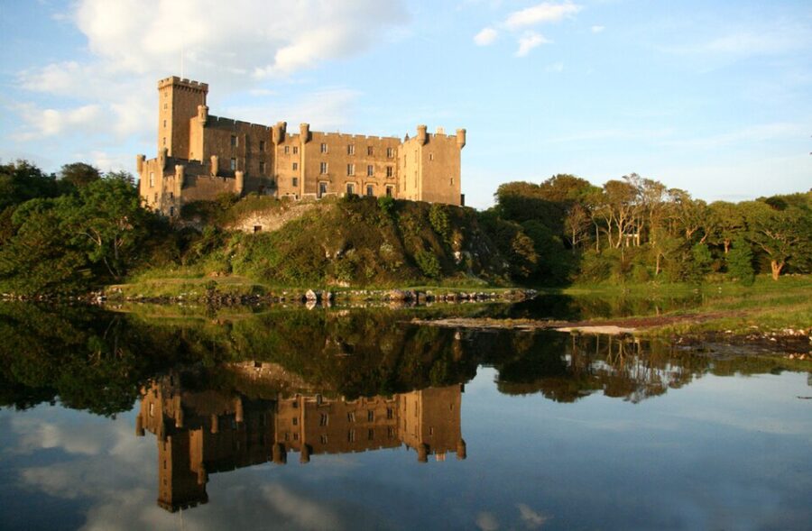 Dunvegan Castle on a hill overlooks a serene lake, set against the scenic backdrop of the Isle of Skye.

