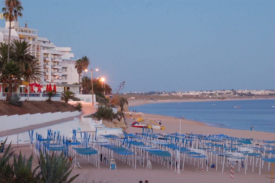 Armacao de Pera Beach resort at twilight with palm trees, red umbrellas, and blue-canopied chairs.