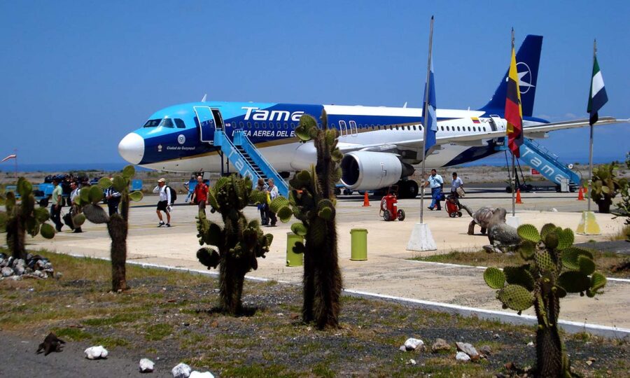 Tame Airlines plane at sunny Ecuador airport with travelers, cacti, and flags in background.