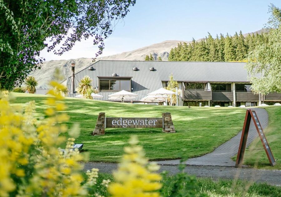 Edgewater mountain lodge resort with green lawn, outdoor patio umbrellas, trees and hills in background.