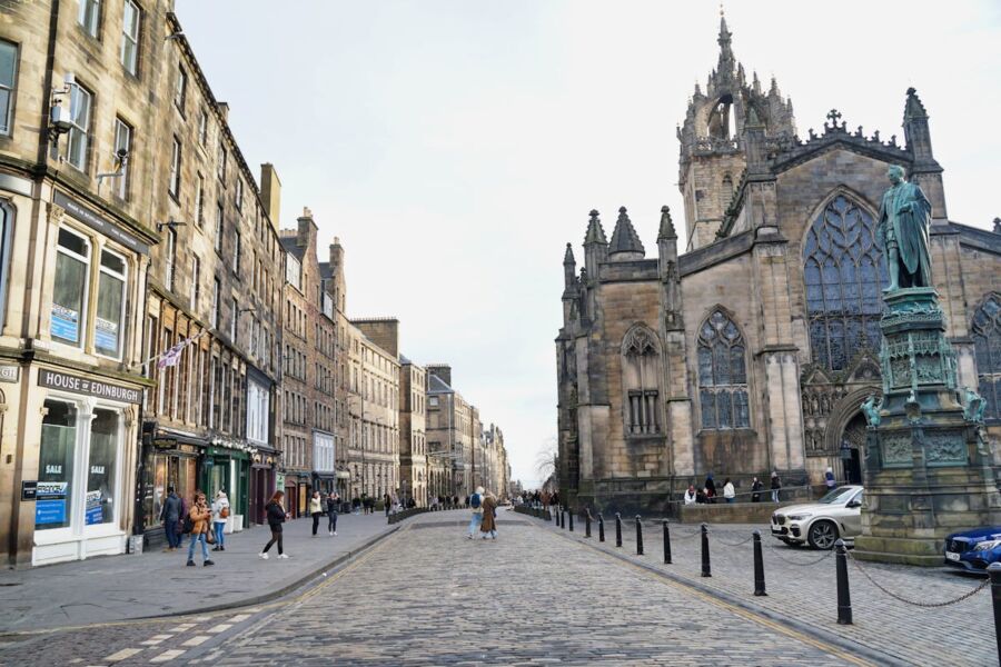 Bustling Edinburgh street scene featuring classic architecture, cobblestones, shops, and a tall spire.