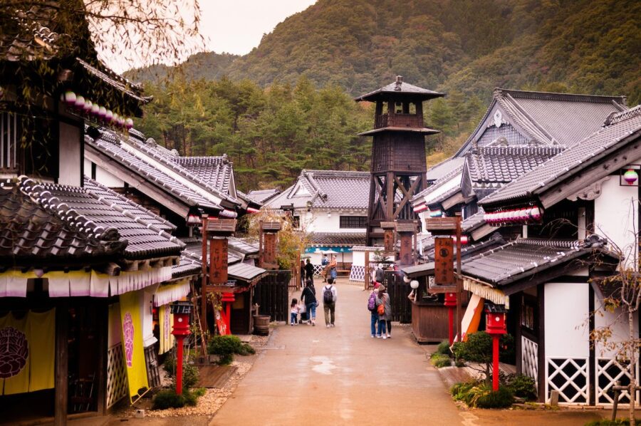 Traditional Edo-period Japanese street with wooden shops, tourists walking, and forested mountains in the background.