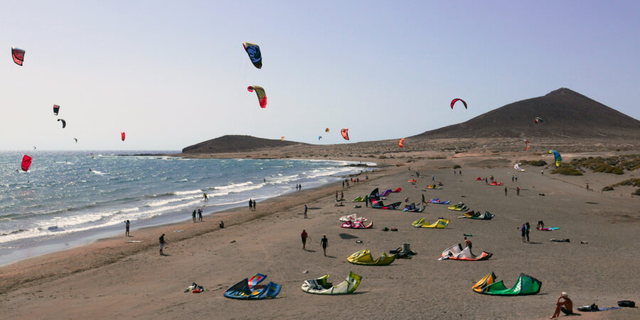 Kitesurfers riding the waves at El Médano beach in Tenerife, with colorful kites against a clear blue sky.
